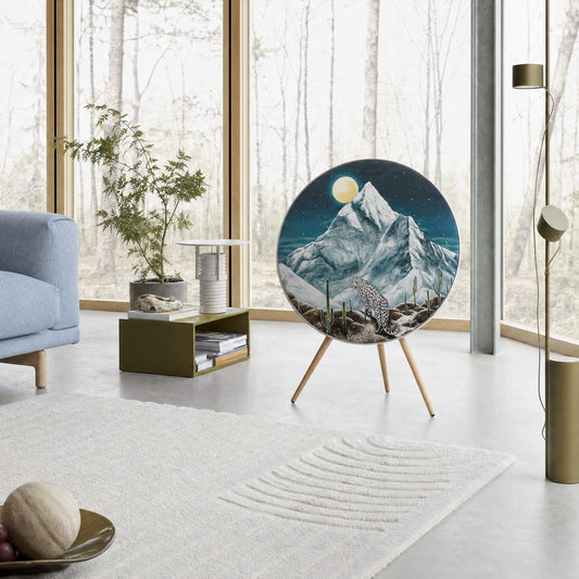 Bang & Olufsen Beosound A9 speaker with the Toward the Moonlight cover by Aira Choi in a bright living room with floor-to-ceiling windows onto bare trees, showing a snow leopard beneath K2 and a full moon