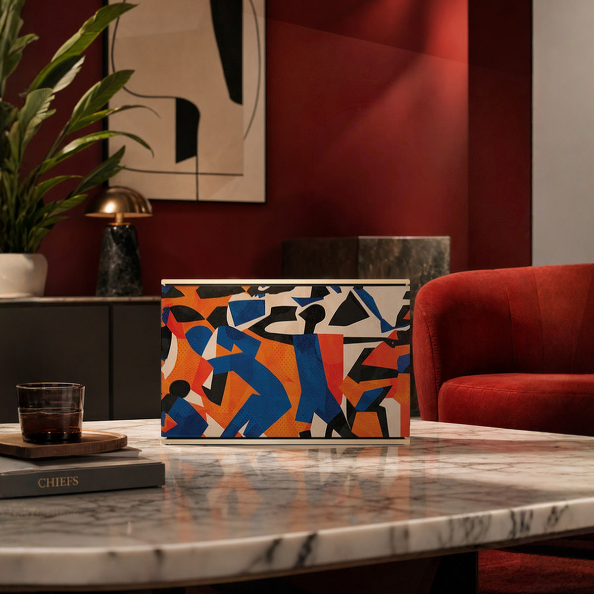 Bang & Olufsen BeoSound Level speaker with Verve cover by Tegrino Studio in a warm residential setting, featuring bold figurative silhouettes in vivid blue, orange and red on marble table beside red velvet armchair with abstract wall art and tropical plant