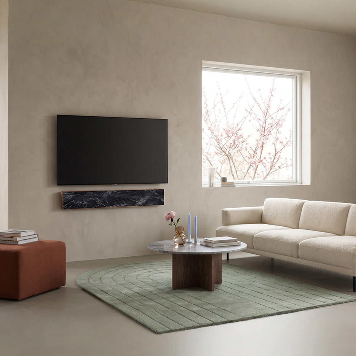 Bang & Olufsen BeoSound Stage soundbar skin in Noir Marquina black marble design with gold veining, mounted below wall TV in warm minimalist living room with terracotta ottoman and sage green rug