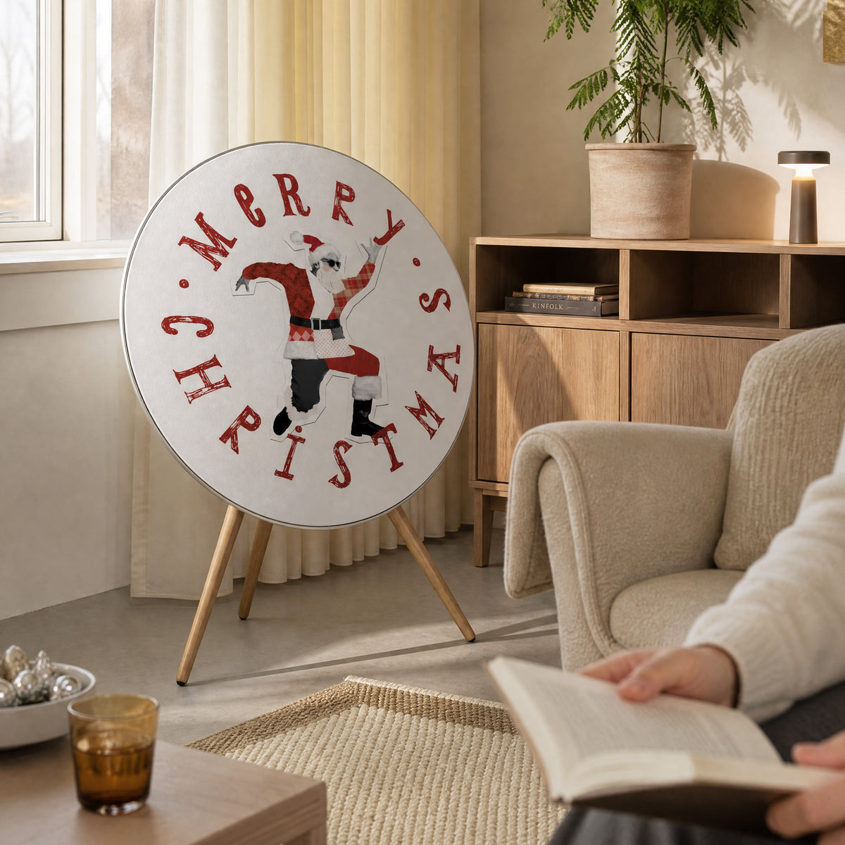 Bang & Olufsen Beosound A9 speaker with the Dancing Santa cover by Tegrino Studio in a sunlit living room beside a reading armchair, showing a collage Santa dancing inside a Merry Christmas ring