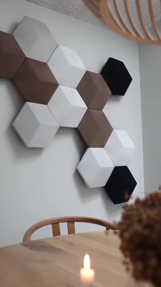 A Bang & Olufsen Beosound Shape system of hexagonal Tegrino felt tiles in white, taupe, sand and black arranged in a honeycomb on a grey wall, above a round oak dining table with candles.