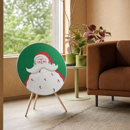 Bang & Olufsen Beosound A9 speaker with the Santa Slopes cover by Tegrino Studio in a wood-panelled living room beside a brown sofa and a window onto green trees, showing tiny skiers on Santa's beard