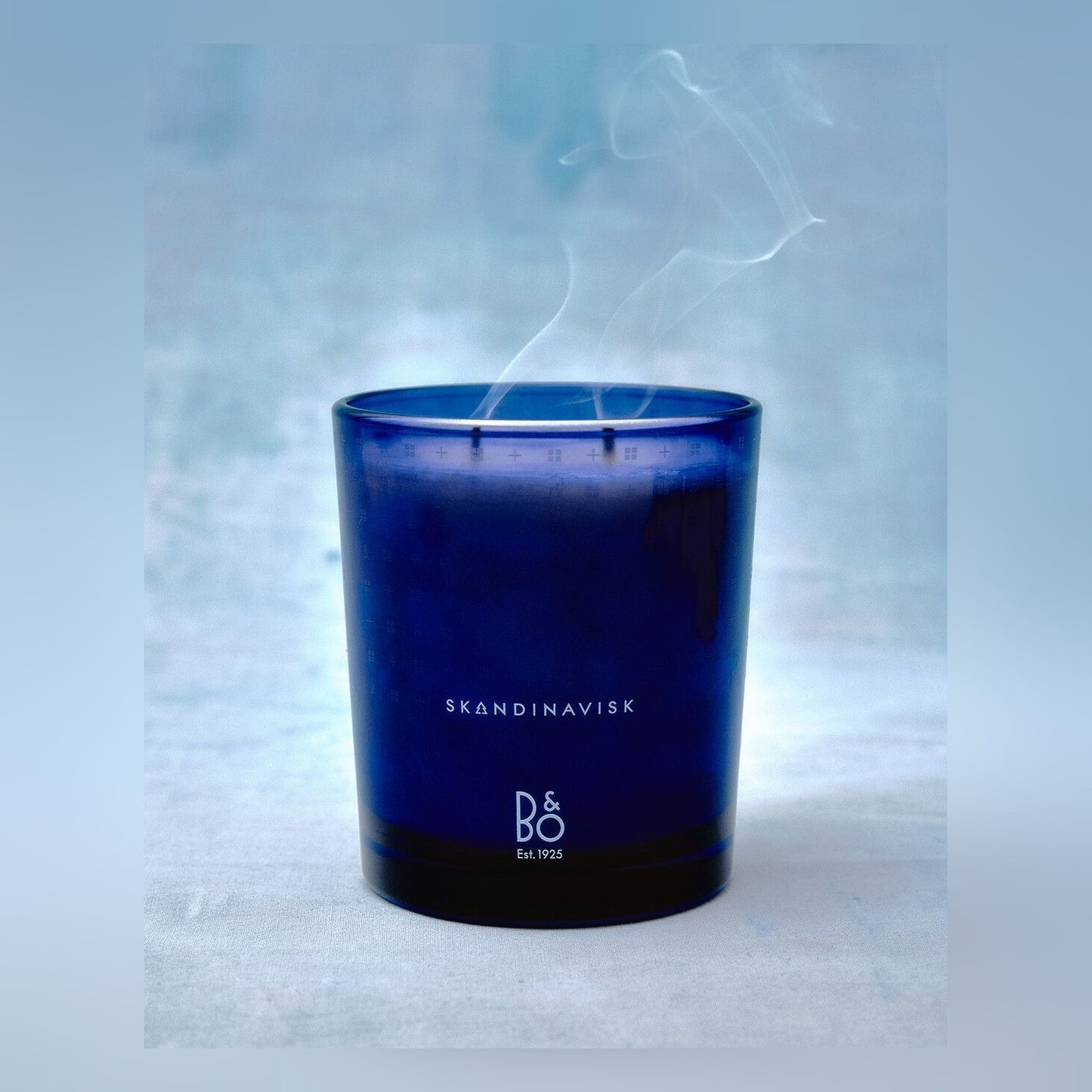 Bang & Olufsen x Skandinavisk centenary limited edition scented candle, lit, in its cobalt blue vessel with both brand marks