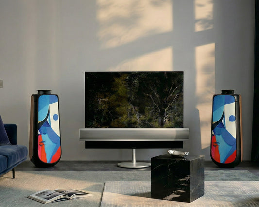Bang & Olufsen BeoLab 50 speaker covers Éclat — Cubist Portrait by Tegrino Studio in a contemporary living space, presenting cubist abstract face in vivid blue, pink and red geometric planes flanking a large TV screen beside navy sofa and black marble cube in sophisticated home cinema setup