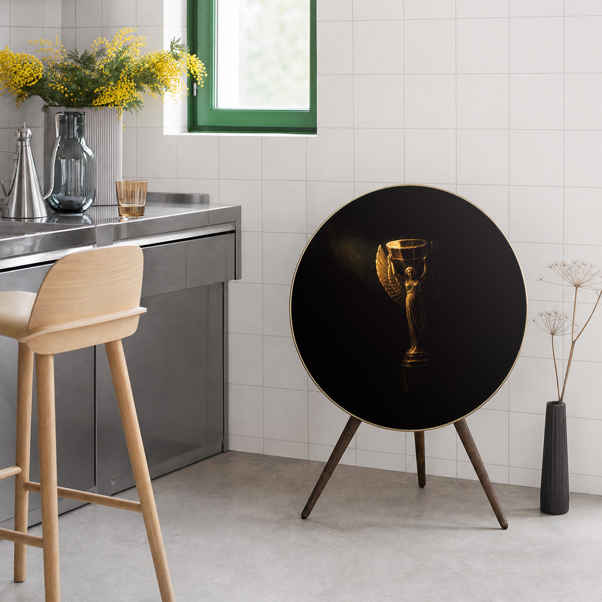 Bang & Olufsen BeoPlay A9 and BeoSound A9 speaker with Nike cover by Tegrino Studio in a carefully styled living area, featuring golden winged victory goddess on deep black ground beside stainless steel kitchen counter with yellow mimosa flowers and oak bar stool