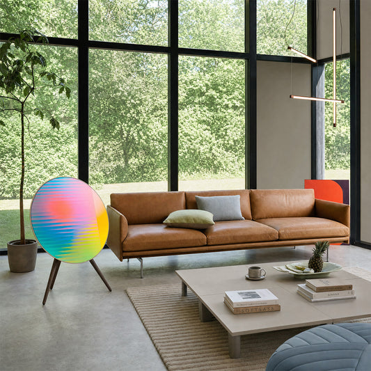 Bang & Olufsen BeoPlay A9 and BeoSound A9 speaker skin Prism by Tegrino Studio in a contemporary living space, featuring full-spectrum gradient scan lines in coral pink, sky blue, teal and yellow beside a tan leather sofa with floor-to-ceiling garden views