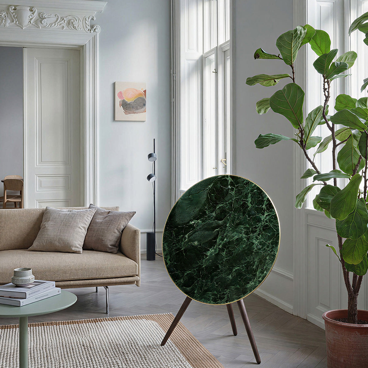 Bang & Olufsen BeoPlay A9 and BeoSound A9 speaker with Verde St. Denis green marble skin displayed in bright European apartment with fiddle leaf fig plant, beige sofa, and ornate crown molding