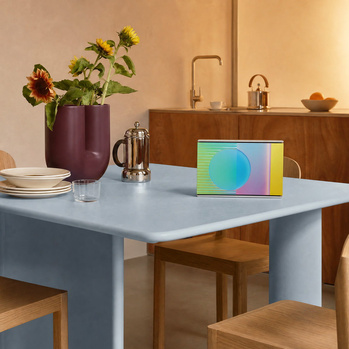 Bang & Olufsen BeoSound Level speaker with Prism cover by Tegrino Studio in a softly lit home setting, presenting holographic sphere with teal, blue and yellow spectrum on powder blue dining table beside sunflowers and french press in warm-toned kitchen interior