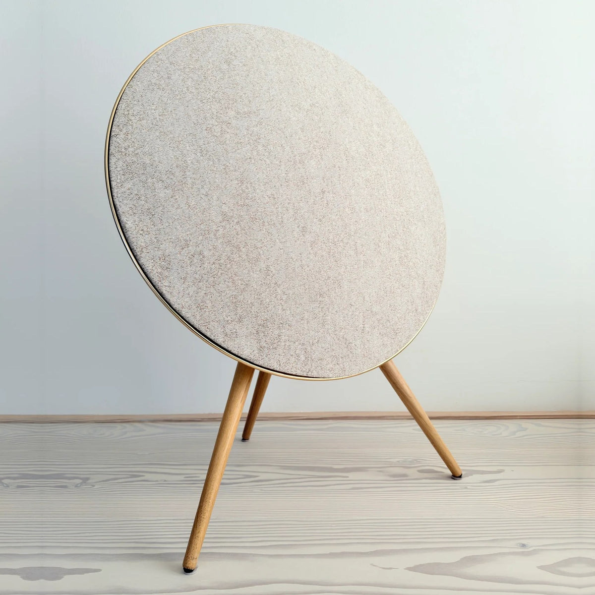 Bang & Olufsen Beoplay A9 and Beosound A9 Kvadrat Woolscape cover Golden by Bang & Olufsen, pale gold knitted wool cover on an A9 with a gold-tone rim and wooden legs, standing on a pale wooden floor against a light wall in daylight